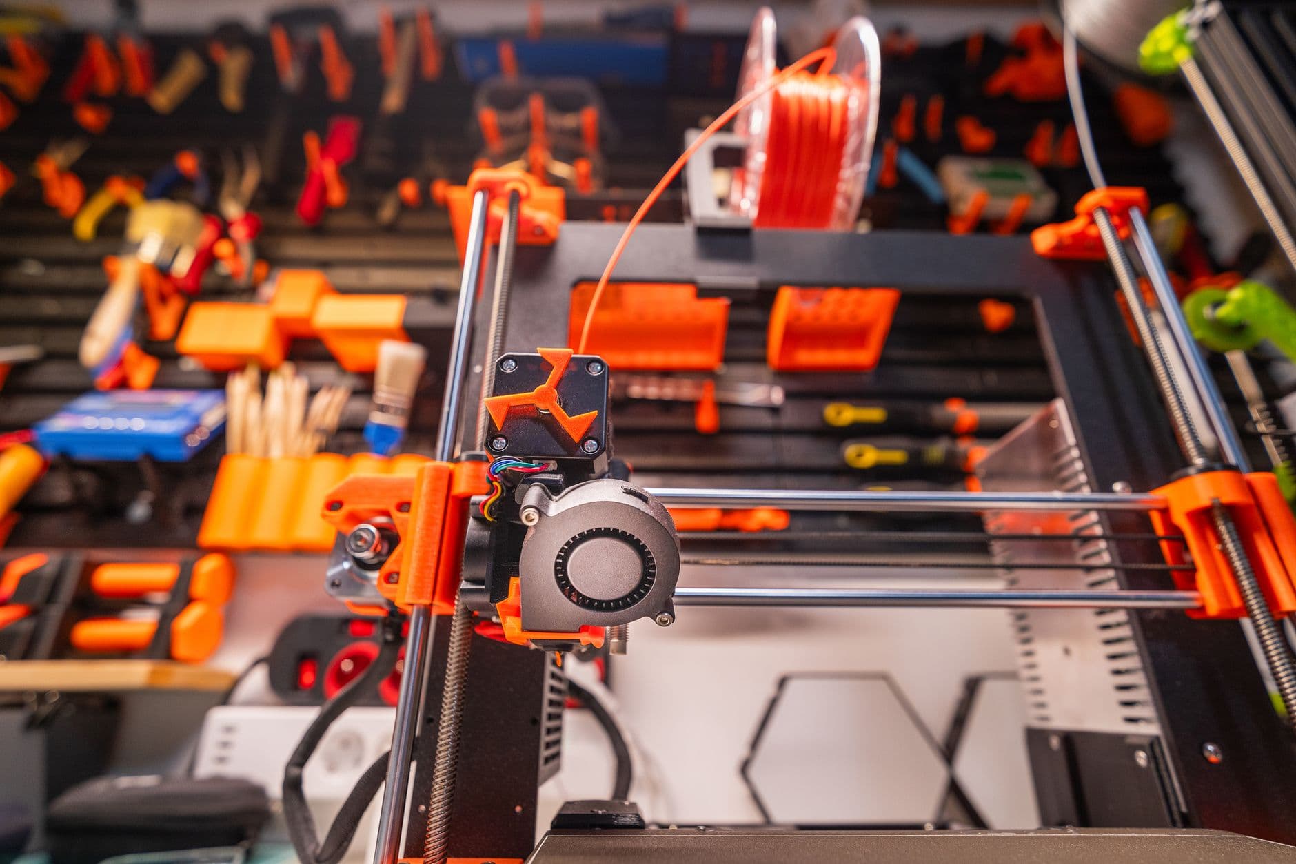 A 3D printer mid-print in a workshop, filament feeding from a spool above a pegboard of tools